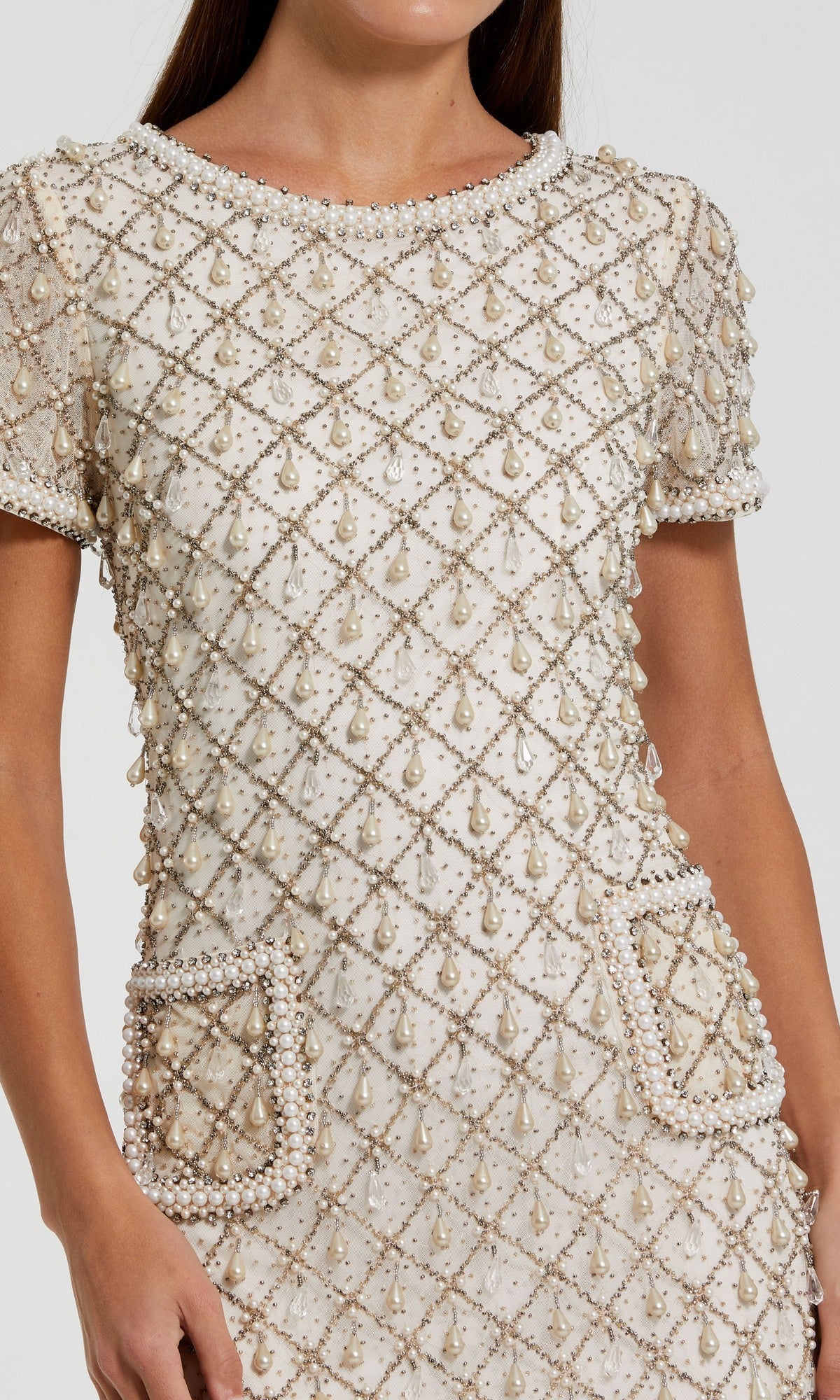 A woman wears the Mac Duggal 10097 short semi-formal dress, featuring cream fabric, short sleeves, a pearl-beaded grid pattern, and two front pockets outlined with pearls.